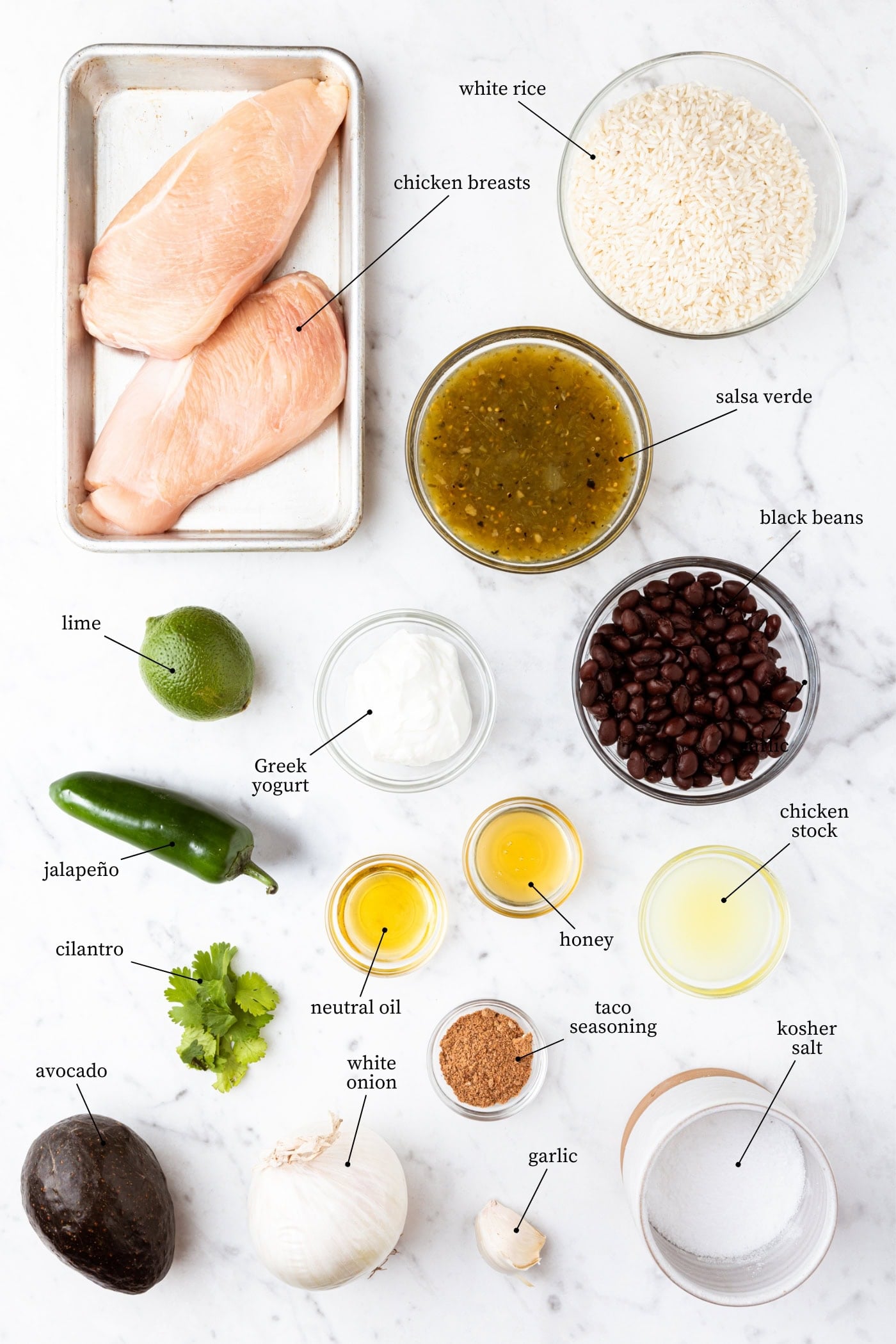 White marble surface with ingredients for chicken rice bowls including rice, beans, chicken, spices, and more.