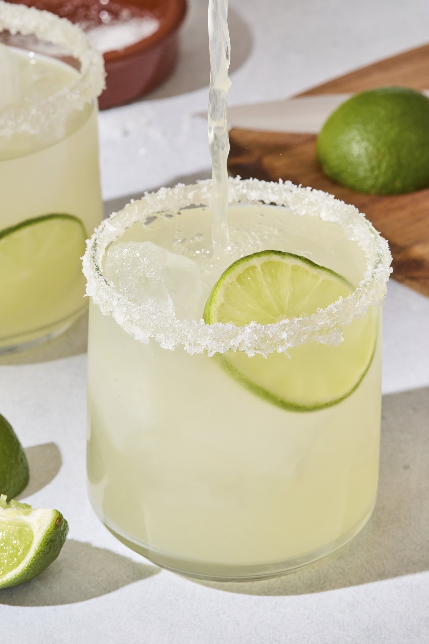 Margarita being poured into a glass with light green margarita already in it with a lime slice.
