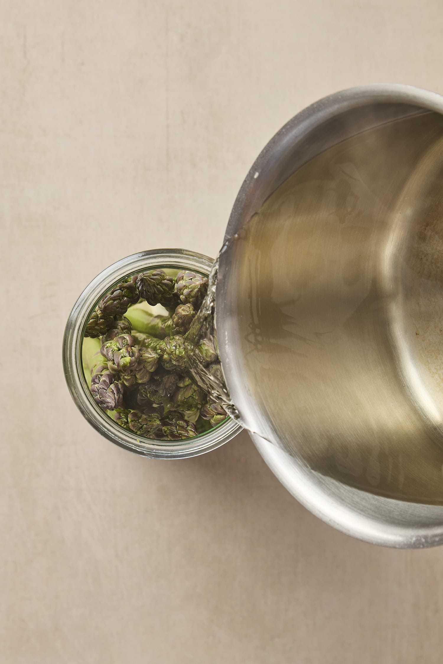 Saucepan with brine pouring into a glass jar filled with asparagus.