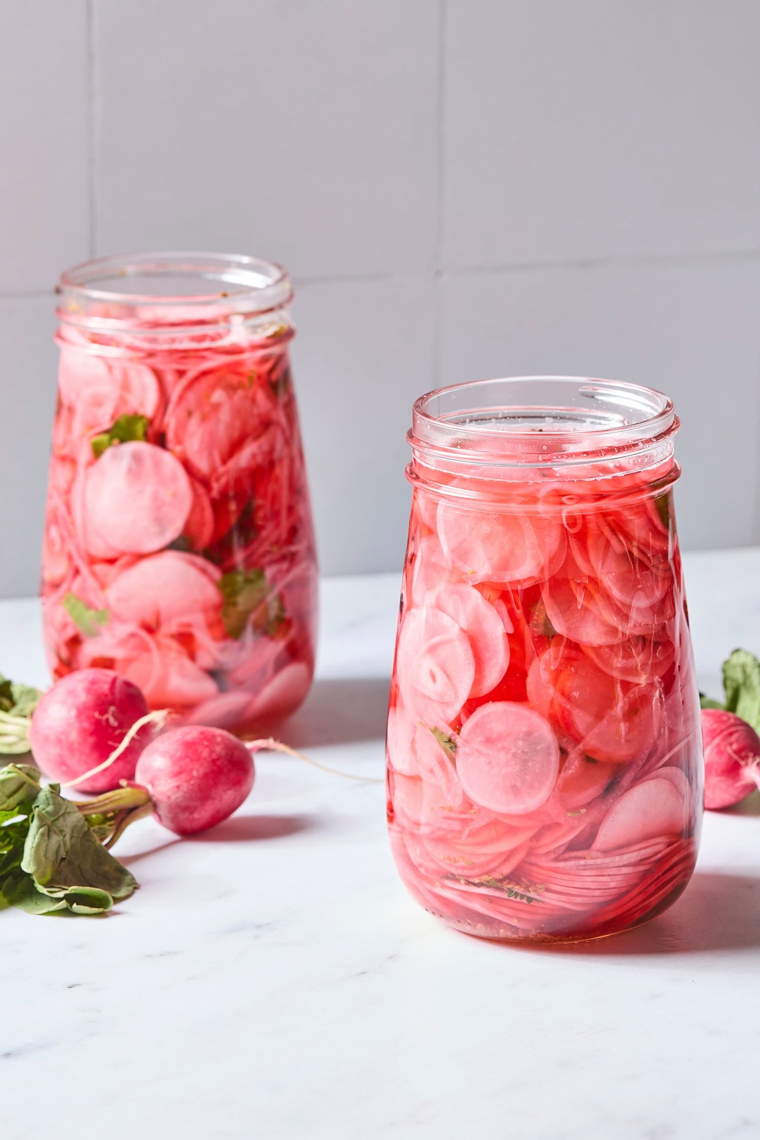 Two glass jars filled with pickled radishes with more radish to the side.