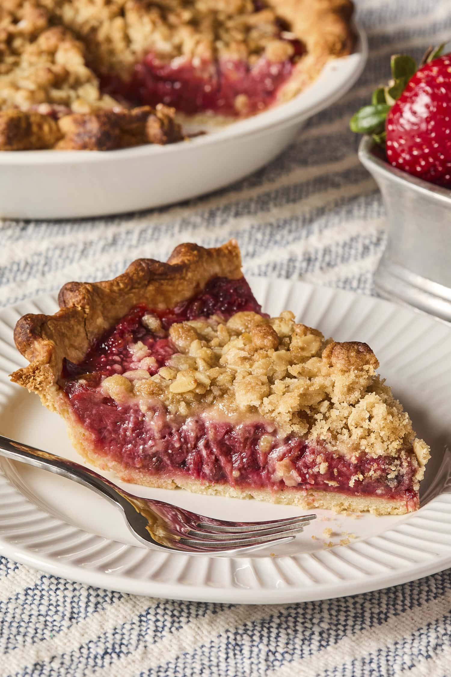 Slice of pie with fork on white plate with more pie and strawberry in background.