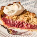 White plate with fork and slice of strawberry rhubarb pie with dollop of whipped cream.