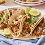 Four chicken tacos with cheese and cilantro on top sitting on a plate.