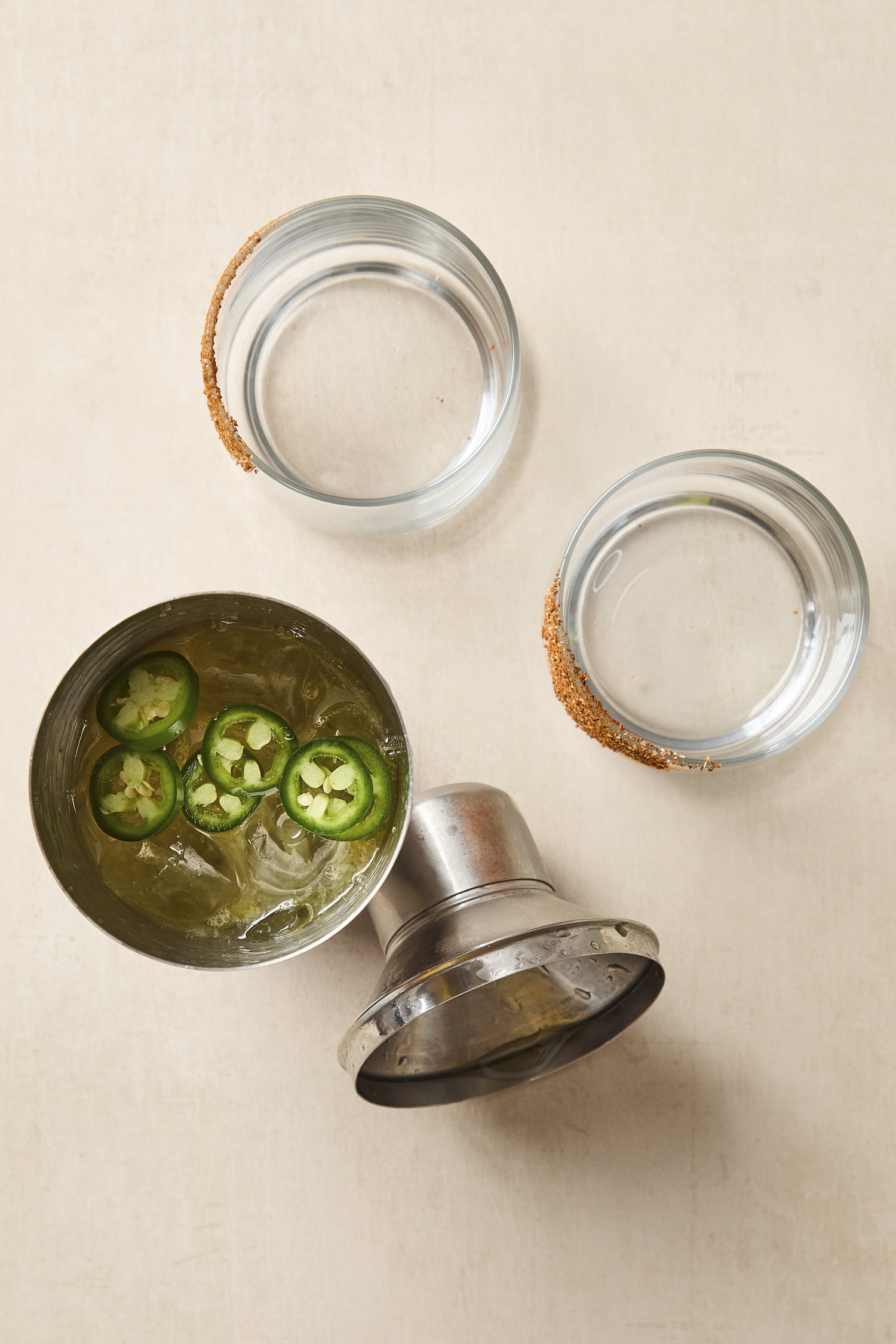 Two glasses with a cocktail shaker filled with prepared margarita before being poured into glass.
