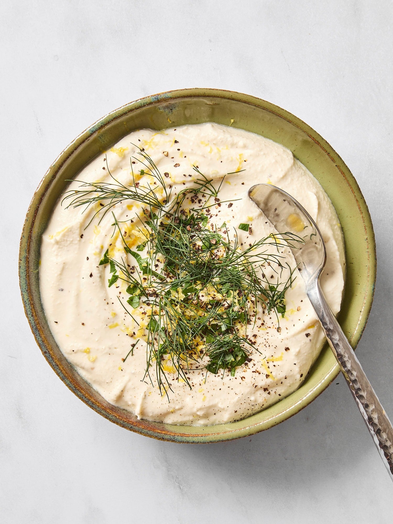 Bowl with feta dip topped with lemon zest, herbs, and olive oil with spoon.