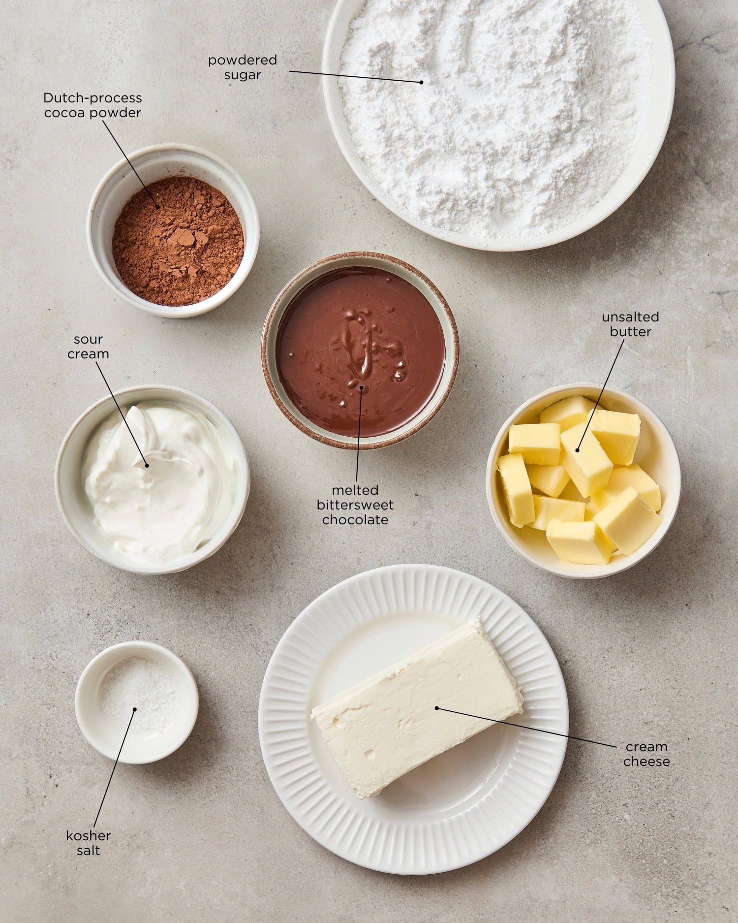 Concrete surface with ingredients needed for chocolate frosting including chocolate, butter, cocoa, and more.