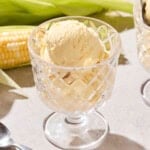 Glass container filled with a serving of ice cream with sweet corn ears in the background.