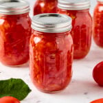A jar of preserved diced tomatoes rests on a marble counter next to tomatoes.