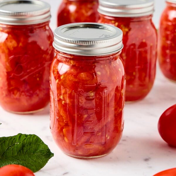A jar of preserved diced tomatoes rests on a marble counter next to tomatoes.