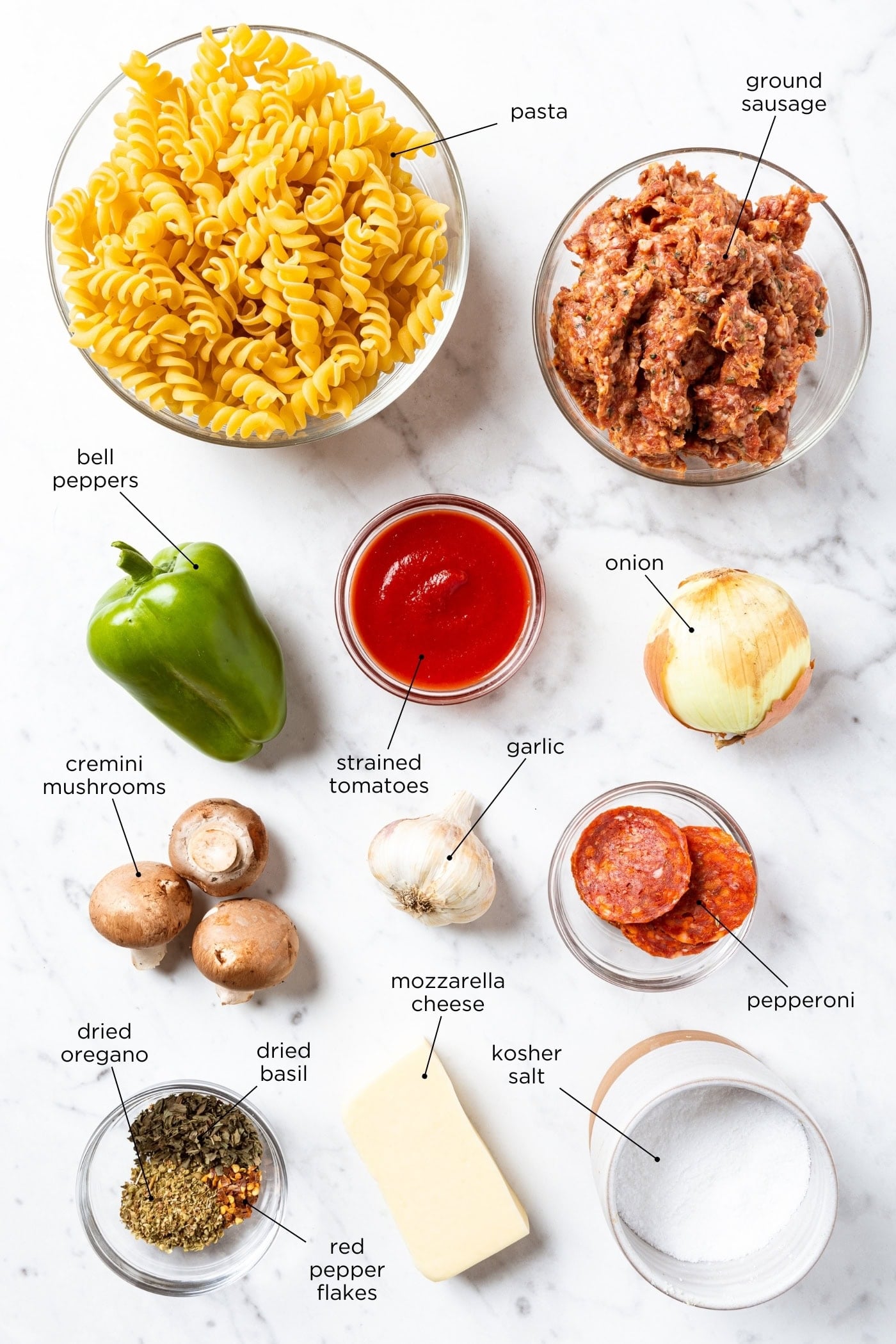 White marble surface with ingredients for pizza casserole including pasta, pepperoni, sauce, veggies, spices, and more.