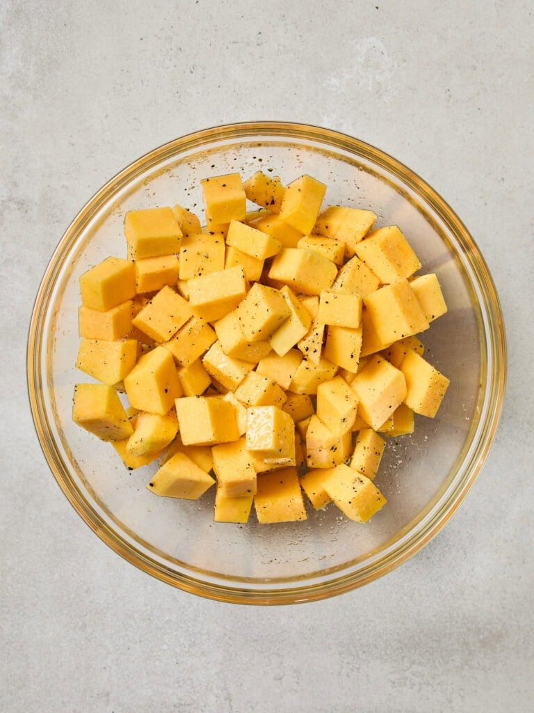Cubed butternut squash being tossed in a bowl with olive oil, black pepper, and salt.