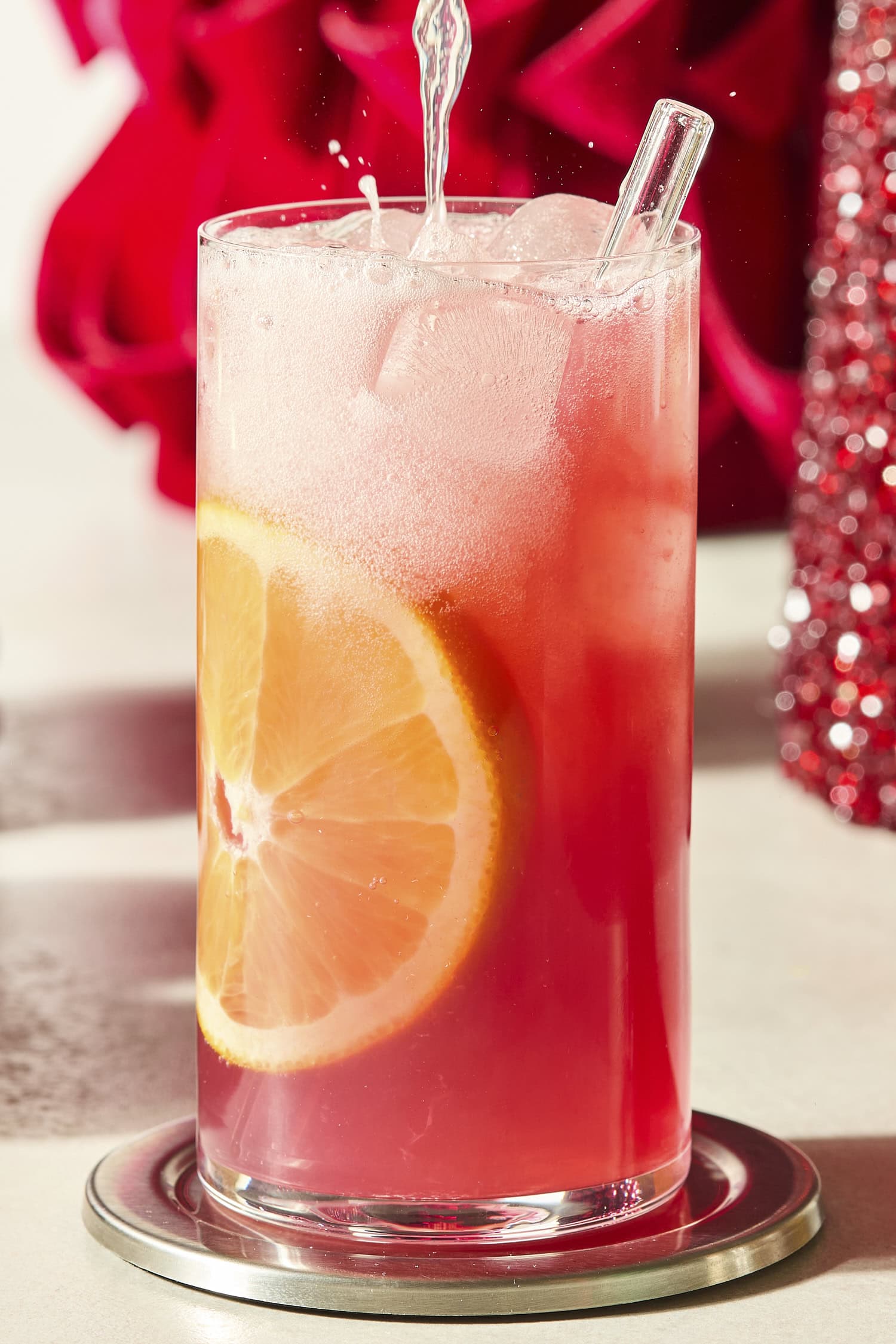 Liquid being poured into a tall cocktail glass filled with cranberry orange mocktail.
