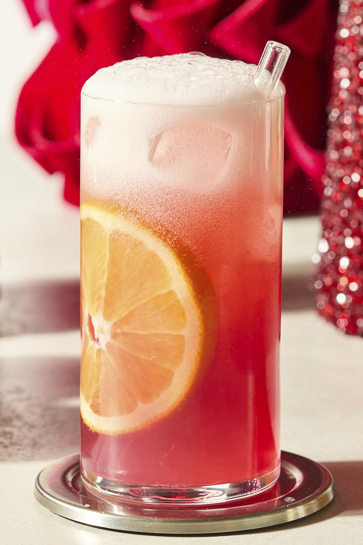 Tall glass with an orange slice down the side and pink color mocktail liquid.