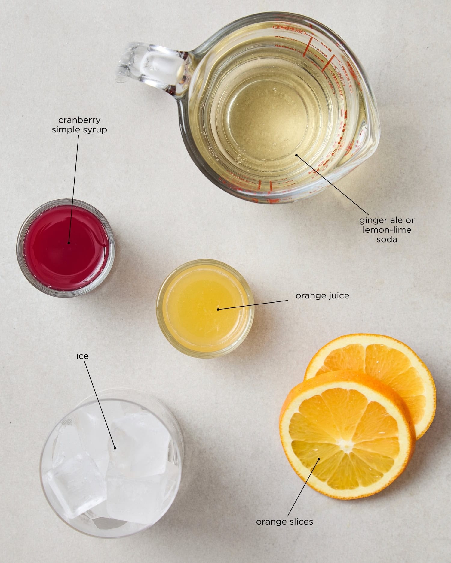 Concrete surface with ingredients for a cranberry mocktail including simple syrup, orange juice, ice, and more.