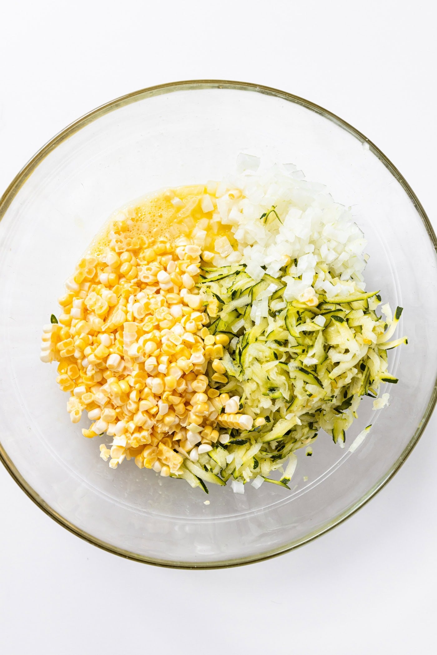 Glass bowl sitting on white countertop filled with onion, corn, and zucchini.