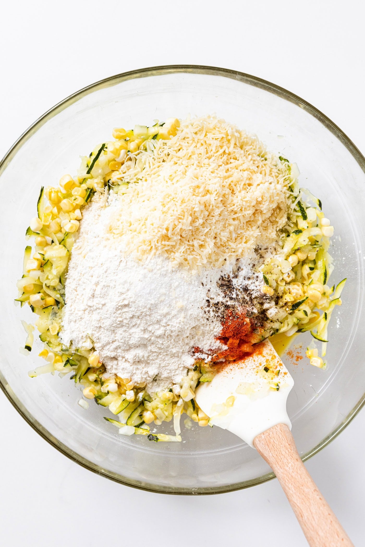 Corn and zucchini mixture sitting in glass bowl topped with flour, Parmesan cheese, and spices.