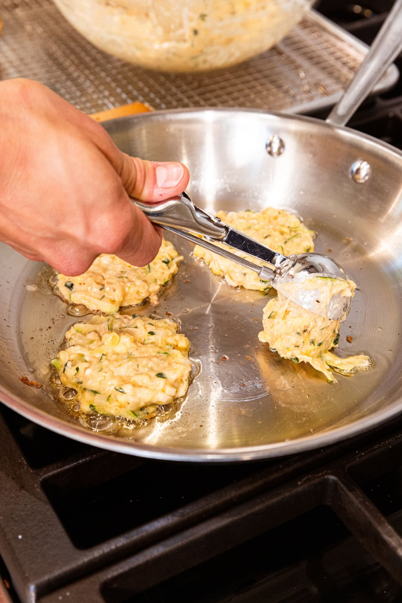 Hand dropping fritter batter from ice cream scoop onto hot metal skillet on stove.
