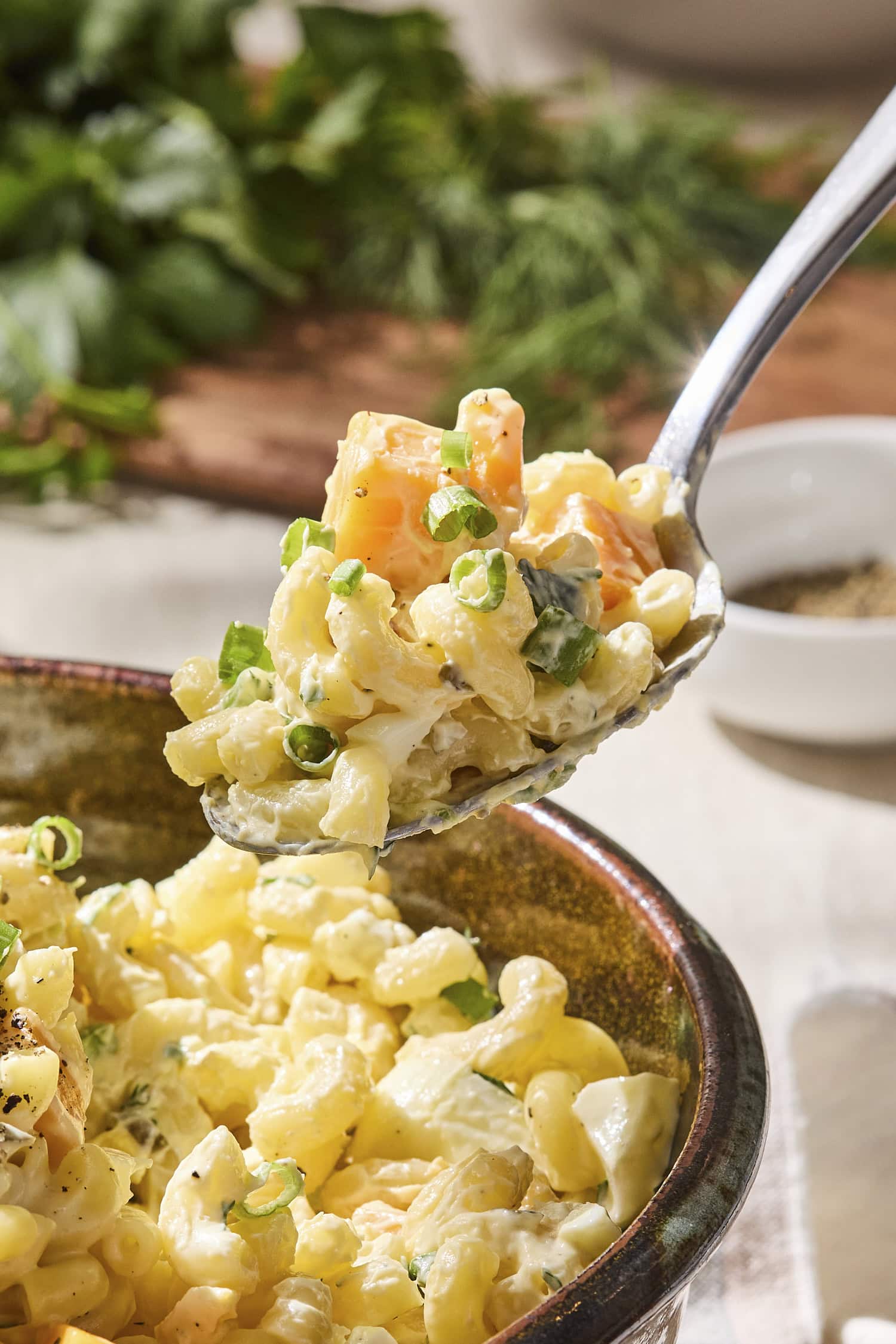 Spoon scooping up serving of macaroni salad with elbow pasta and cheese cubes.
