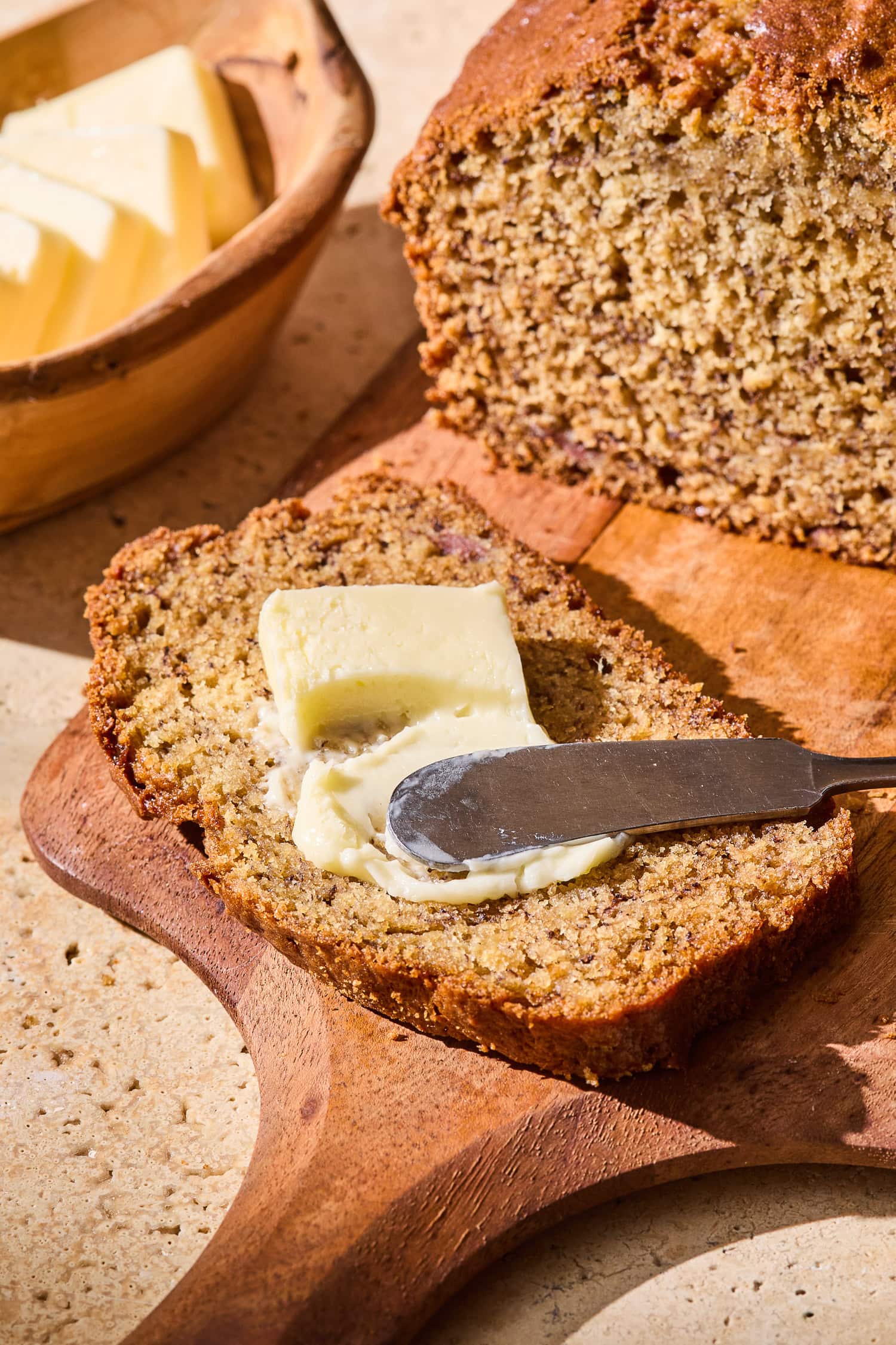A slice of classic banana bread getting a slice of butter spread overtop.