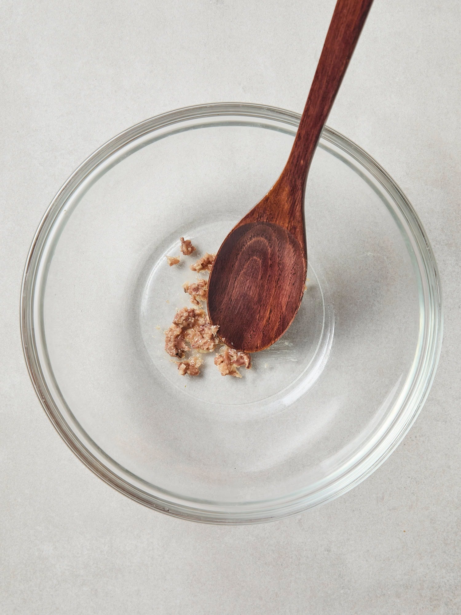 Glass bowl with wooden spoon mashing anchovies and garlic.