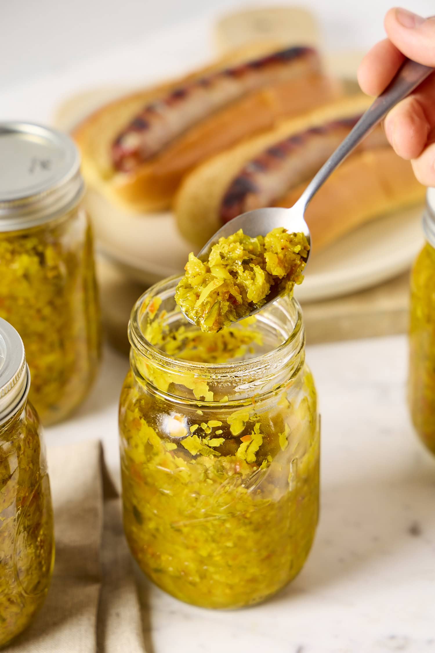 Spoonful of zucchini relish from a glass jar with brats in background.
